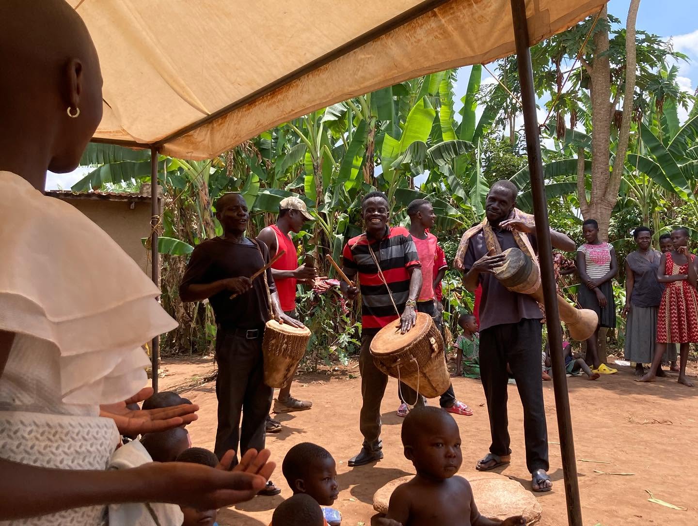 Men play traditional drums outdoors while children and families watch under a canopy