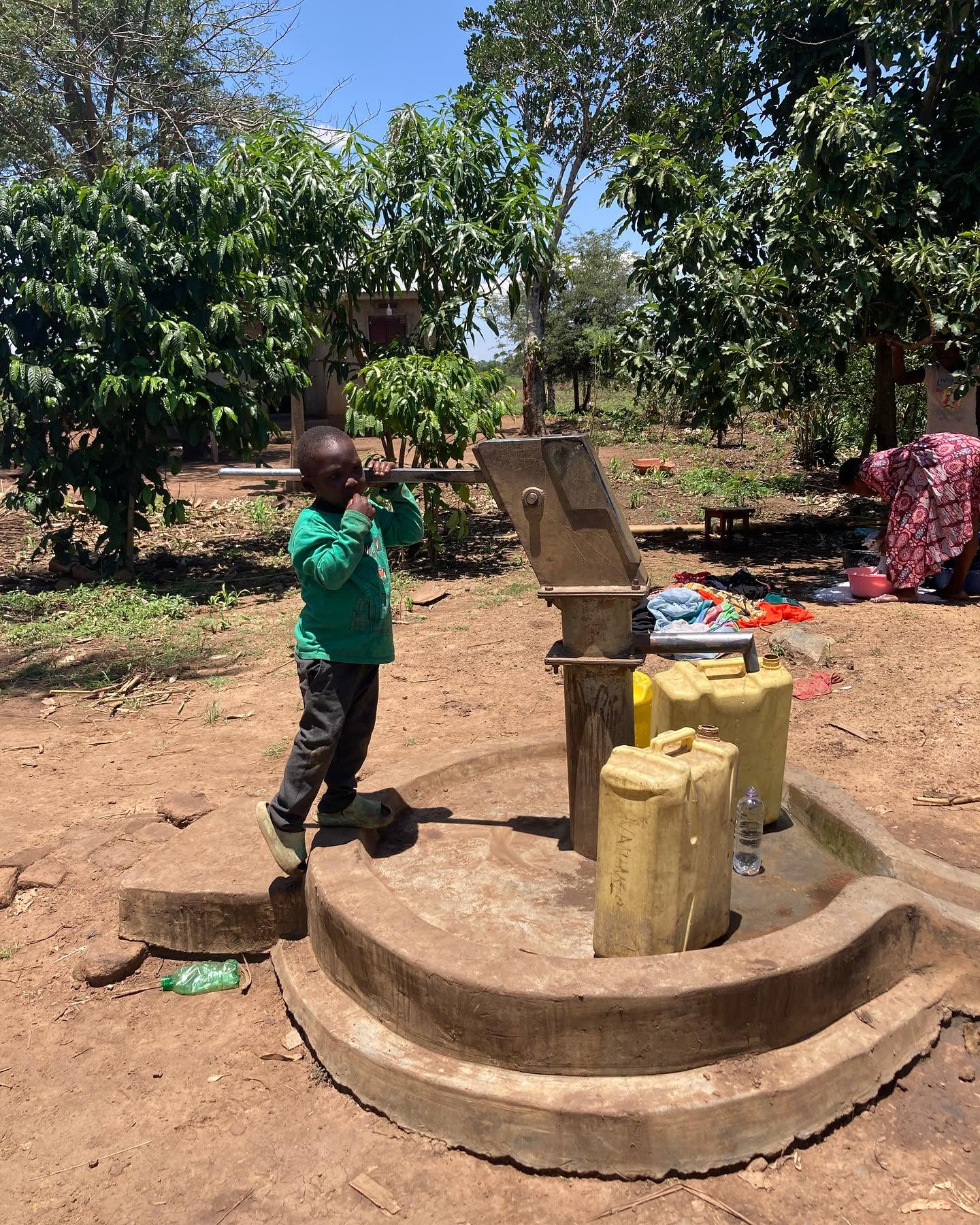 A child draws water from a hand pump while containers wait nearby