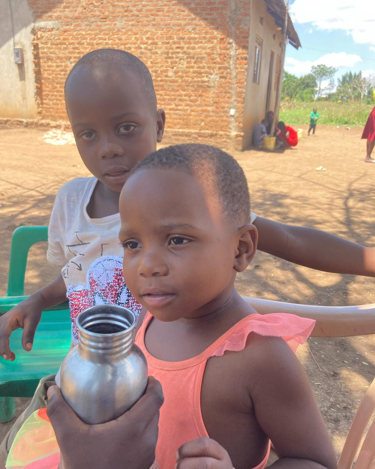 Two young children sit together outdoors, one holding a metal water bottle