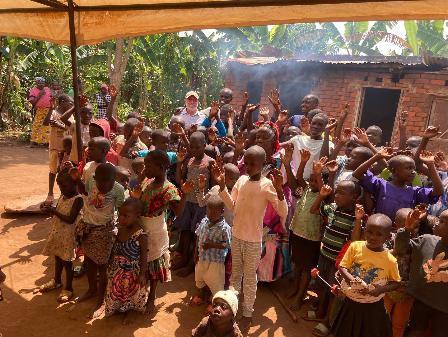 A large group of children and adults gather under a canopy with raised hands