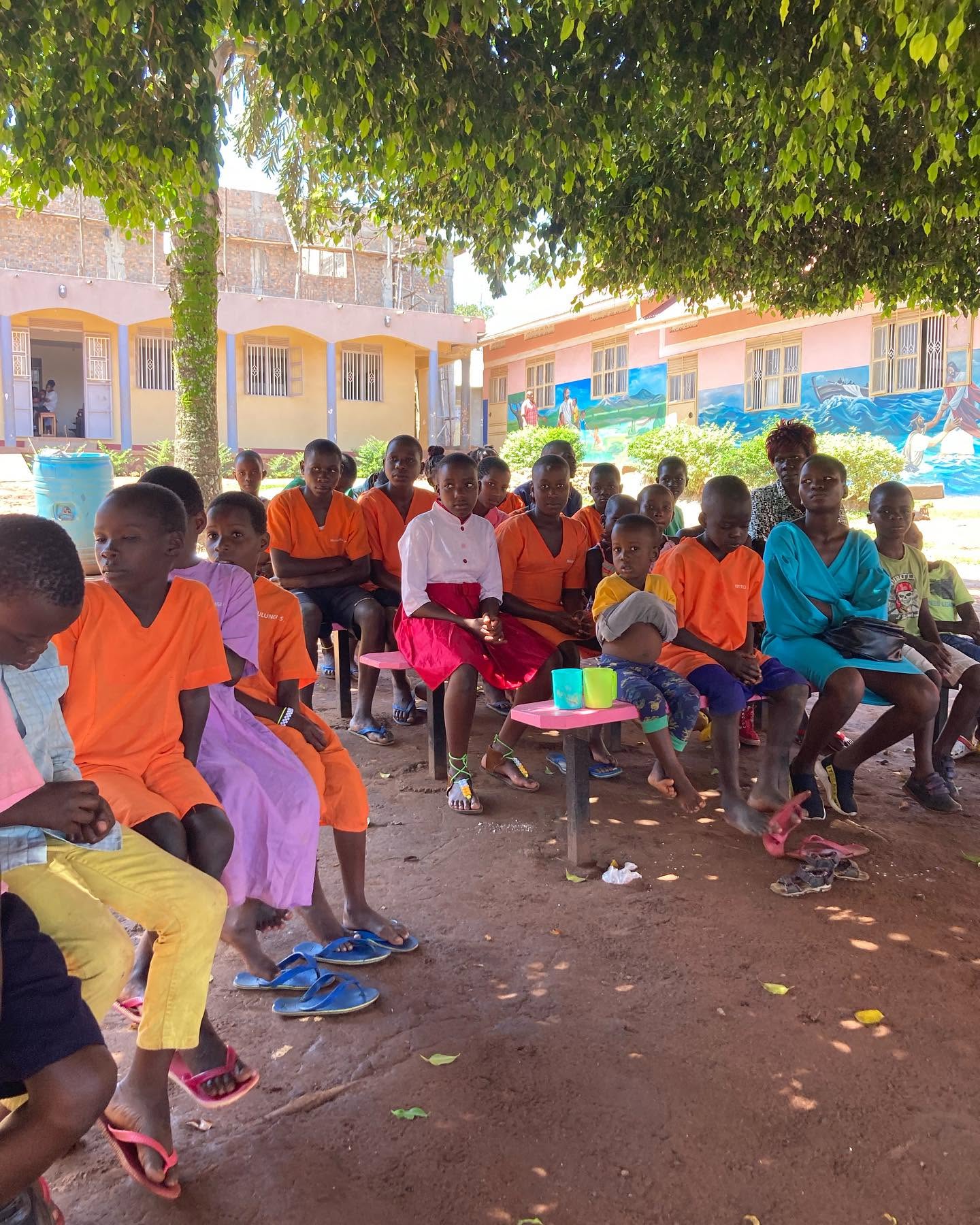 Students in bright uniforms gather in the shade of a tree at school